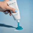 Load image into Gallery viewer, A hand with light blue nail polish squeezes a white tube of Blue M toothpaste, with bright blue toothpaste gel squirting onto a light blue surface.
