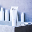 Load image into Gallery viewer, A white tube of blue m toothpaste stands on a concrete ledge in a bathroom setting, with blurred toothpaste bottles in the background.

